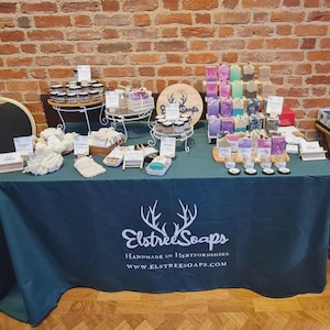May include: A stall showcasing handmade soaps and skincare items. The products are arranged on a green tablecloth, with a variety of colourful soaps, jars of creams, and other items. The tablecloth displays the text "Elstree Soaps" and the website address.