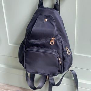 May include: Black nylon backpack with gold-coloured zips and hardware. The small backpack has a top handle, adjustable straps, and a front pocket. The bag is hanging on a light green door.