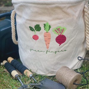May include: A beige canvas gardening bucket with embroidered vegetables and the text "green fingers." The bucket has rope handles and sits on grass next to gardening tools and twine.