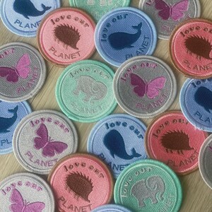 May include: A collection of embroidered patches with the text "love our planet." The patches feature various animal designs, including a whale, butterfly, elephant, and hedgehog, in shades of blue, pink, gray, and green.