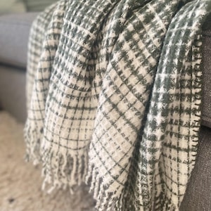 Könnte beinhalten: Eine weiche, gewebte Decke mit grün-weißem Karomuster. Die Decke ist über ein graues Sofa drapiert und hat einen Fransenbesatz am Rand. Die Textur wirkt flauschig und gemütlich, perfekt, um einem Wohnraum Wärme zu verleihen.