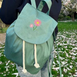 May include: A teal backpack with a flap and drawstring closure. The flap features an embroidered design of two pink and yellow flowers with green stems. The backpack has white rope drawstrings and teal straps. The background shows a person wearing the backpack.