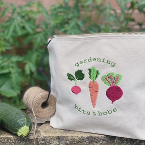 May include: Off-white canvas zippered pouch with embroidered vegetables: radish, carrot, and beet. Text reads "gardening bits & bobs" in green. A zucchini and twine are in the foreground. Ideal for gardeners.