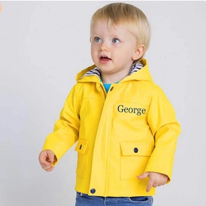 Puede incluir: Una gabardina amarilla con capucha y una franja azul en el interior de la capucha. La gabardina tiene una cremallera y un cierre de botón. La gabardina tiene un bolsillo en la parte delantera con un cierre de botón. La gabardina tiene el nombre "George" bordado en la parte delantera.