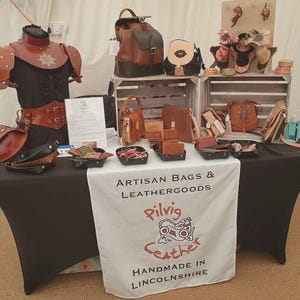 May include: A stall showcasing artisan leather bags and accessories in shades of brown and black. Items include backpacks, purses, wallets, and belts. The table is draped with a white cloth bearing the text "ARTISAN BAGS & LEATHERGOODS" and "Pilvig Leather Handmade in Lincolnshire."