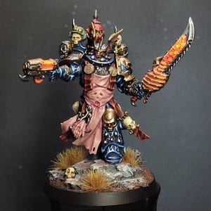 May include: A miniature figurine of a warrior in dark blue and red armor, holding a glowing orange pistol and a serrated sword. The figure is adorned with skulls and stands on a rocky base with dry grass.