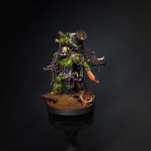 May include: A detailed miniature figure of a warrior in green and purple armor, carrying a large weapon on its back. The figure stands on a brown base with a skull and grass. The weapon has a metallic finish with orange accents.