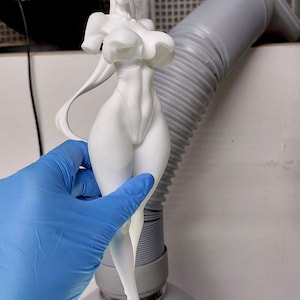 May include: A white 3D printed figurine of a female figure with long hair and pointed ears. The figure is wearing a one-piece outfit with a high neckline and a low-cut back. The figure is posed with one hand on her hip and the other arm extended.