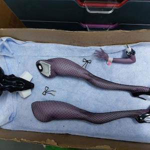 May include: A set of doll parts including a torso, legs, and arms. The doll has black fishnet stockings and black heels. The doll parts are laid out on a blue fabric surface.
