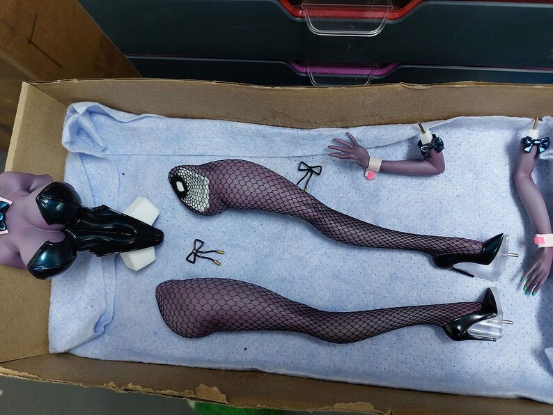 May include: A set of doll parts including a torso, legs, and arms. The doll has black fishnet stockings and black heels. The doll parts are laid out on a blue fabric surface.