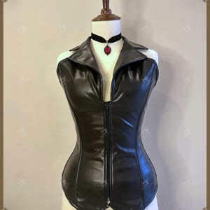 May include: A black faux leather corset with a zipper closure and a collared neckline. A black choker necklace with a red pendant is worn around the neck. The corset is displayed on a mannequin.