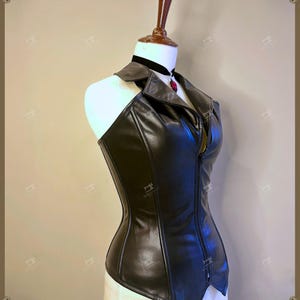 May include: A black faux leather corset with a halter neckline and a front zipper. The corset is displayed on a mannequin with a black choker and a red pendant. The garment has a tailored fit and a structured design.