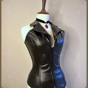 May include: A black faux leather corset with a collared neckline and a front zipper. The corset is displayed on a mannequin and is paired with a black velvet choker featuring a red pendant.