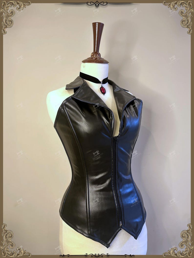May include: A black faux leather corset with a collared neckline and a front zipper. The corset is displayed on a mannequin and is paired with a black velvet choker featuring a red pendant.