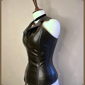 May include: A black faux leather corset with a zipper closure and a collar. The corset is displayed on a mannequin and features a fitted silhouette. A black choker with a red pendant is visible.