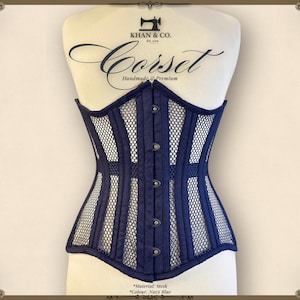 May include: A navy blue corset made of mesh fabric with a busk closure and metal eyelets. The corset has a structured design with vertical panels and a fitted silhouette. "KHAN & CO. Corset Handmade Premium" is printed on the top of the corset.