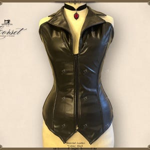 May include: Black leather corset with a collared neckline and a front zipper closure. The corset has a fitted silhouette and is displayed on a mannequin. A black choker with a red pendant is worn around the mannequin's neck. The corset is handmade.