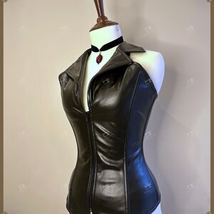 May include: A black faux leather corset top with a zipper closure, displayed on a mannequin. The top features a collar and is paired with a black velvet choker with a red pendant. The corset is designed to accentuate the waist.
