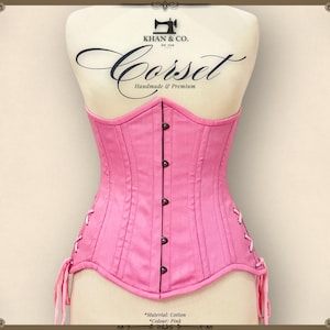 Può includere: Un corsetto in cotone rosa con dettagli di cucitura neri e una fila di bottoni sul davanti. Il corsetto è fatto a mano e presenta un'etichetta che recita "Khan & Co. Est. 2019 Corset Fatto a mano & Premium".