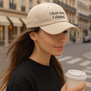 Pode incluir: Boné de beisebol bege com o texto "I dont chase, I channel" em preto. O boné é usado por uma pessoa vestindo um moletom preto e segurando uma xícara de café branca. O fundo mostra uma rua da cidade.