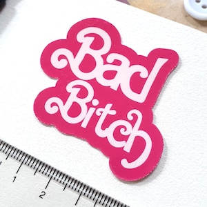Op de afbeelding: Een felroze sticker met de tekst "Bad Bitch" in witte letters. De sticker ligt op een wit oppervlak, naast een transparante liniaal, een zwarte schaar en een witte knoop. Een roze en bruin patroonpapier en papier met sterrenprint zijn op de achtergrond.