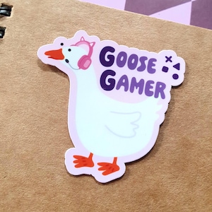Könnte beinhalten: Ein Aufkleber mit einer weißen Gans mit rosa Kopfhörern und dem lila Schriftzug "GOOSE GAMER". Die Gans hat orangefarbene Füße und einen rosa Schnabel. Der Aufkleber befindet sich auf einem braunen Notizbuch.