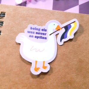 Puede incluir: Una pegatina con un ganso blanco con patas y pico naranjas, sosteniendo una bandera del orgullo no binario. La pegatina tiene el texto "being cis was never an option" en morado. La pegatina está sobre un cuaderno marrón.