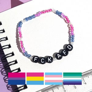 Può includere: Un braccialetto di perline con una combinazione di colori bi-orgoglio, con perline rosa, viola e blu. Il braccialetto ha perline di lettere nere che compongono "FCK AFD". L'immagine include diversi disegni di bandiere dell'orgoglio.