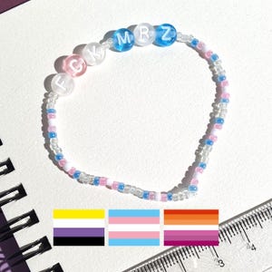 Op de afbeelding: Een kralenarmband met de letters FCKMRZ in witte en blauwe kralen. De armband is gemaakt van kleine, kleurrijke kralen in roze, blauw en transparant. Drie LGBTQ+-vlaggen worden eronder weergegeven.