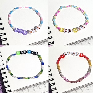 Könnte beinhalten: Vier Armbänder mit Perlen, die den Schriftzug "FCK NZS" bilden. Jedes Armband hat ein anderes Farbschema, darunter blaue, rosa, gelbe, grüne und rote Perlen. Die Armbänder liegen auf einer weißen Oberfläche mit einem Lineal.