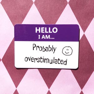 Op de afbeelding: Een paars en wit naamlabel met de tekst "HELLO I AM... Probably overstimulated" en een smiley. De achtergrond is een roze en wit ruitpatroon.