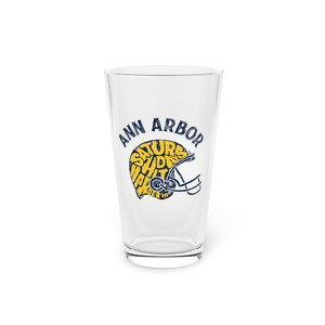 Könnte beinhalten: Ein klares Pintglas mit den Worten "ANN ARBOR" in Marineblau über einer gelben und marineblauen Footballhelm-Grafik. Die Helm-Grafik enthält die Worte "SATURDAY SHIRT PARTY" in Gelb.