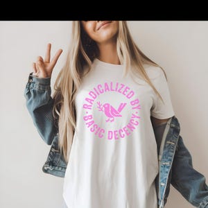 以下が含まれることがあります： ピンク色のグラフィックが特徴の白いTシャツ。円形のデザインで「RADICALIZED BY BASIC DECENCY」と書かれています。グラフィックには、枝を持ったピンク色の鳥が含まれています。画像内の人物はデニムジャケットを着ています。