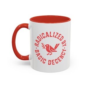May include: White ceramic mug with a red interior and handle. The mug features the text "RADICALIZED BY BASIC DECENCY" in red, encircling a red bird graphic holding a sprig.