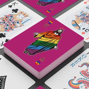 May include: A deck of playing cards with a magenta back. The face card features a rainbow-colored Jesus illustration. Other cards visible include a King of Clubs and a Joker card. The deck is stacked on a white surface.