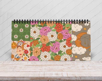 Könnte beinhalten: Ein spiralgebundenes Notizbuch mit einer Collage von Retro-Designs. Die Designs umfassen florale Muster, eine himmlische Mond- und Wolkenillustration und andere skurrile Grafiken. Das Notizbuch liegt auf einer Holzoberfläche.
