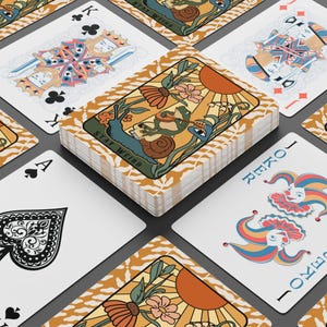 May include: A deck of playing cards with a whimsical design. The cards feature illustrations of a snail, frog, and sun, with the words "Stay Weird" on the card backs. The face cards have traditional suits and face values.