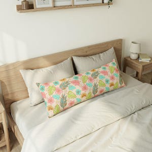 May include: A long body pillow with a tropical floral pattern. The pillow features pink, blue, green, and yellow flowers and leaves on a light peach background. The pillow is on a bed with neutral-colored bedding.