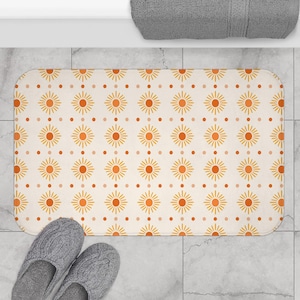 May include: A rectangular bath mat featuring a repeating pattern of orange sunburst icons and small orange dots on a cream background. The mat has rounded corners and is positioned on a light gray tiled floor with a gray towel visible above it.
