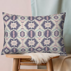 May include: Rectangular pillow with a geometric pattern in shades of purple, pink, and light blue on a cream background. The pillow is placed on a wooden stool with a white fur throw.