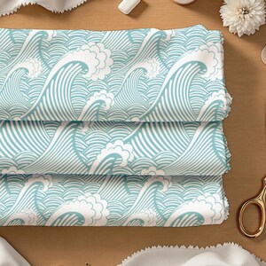 May include: A folded piece of fabric featuring a repeating pattern of stylized ocean waves in light blue and white. The design consists of curved lines and foam crests, creating a continuous graphic print across the textile surface.