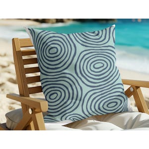 Peut inclure: Coussin carré avec un fond bleu clair et un motif de cercles concentriques bleu foncé. Le coussin est posé sur une chaise en bois avec un coussin blanc, sur une plage.