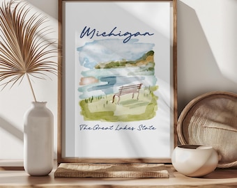 May include: A framed watercolor print featuring a Michigan landscape with a bench, water, and hills. The print has the word "Michigan" in blue script and the text "The Great Lakes State" below the image. The frame is made of wood.