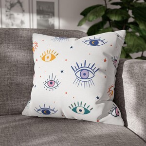 May include: Square white cushion featuring a repeating pattern of colourful eyes, stars, and dots. The eyes are in various colours, including blue, pink, yellow, and green. The cushion is on a grey sofa.