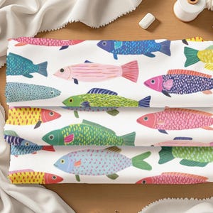 May include: A stack of three folded fabric pieces featuring a repeating pattern of colorful fish on a white background. The fish are illustrated in various shades of blue, pink, yellow, and green with distinct scale textures and fin details.