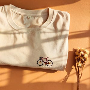 Könnte beinhalten: Ein gefaltetes, cremefarbenes T-Shirt mit einer kleinen, gestickten Fahrradzeichnung in Rot und Schwarz. Das T-Shirt, Größe L, liegt auf einer orangefarbenen Oberfläche, begleitet von Trockenblumen.