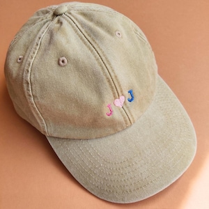 Könnte beinhalten: Beige Baseballkappe mit gebogenem Schirm. Die Kappe hat gestickte Buchstaben "J" in Pink, ein pinkfarbenes Herz und ein blaues "J". Die Kappe hat ein klassisches Sechs-Panel-Design mit einem oberen Knopf und einem verstellbaren Riemen.