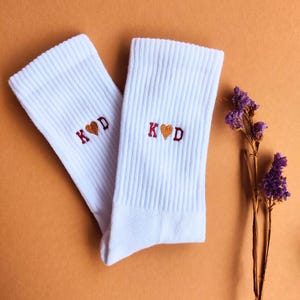 Könnte beinhalten: Ein Paar weiße gerippte Socken mit den gestickten Initialen "K ❤️ D" in einem Herzdesign. Die Socken werden auf einem orangefarbenen Hintergrund mit einem Zweig lila Blumen präsentiert.