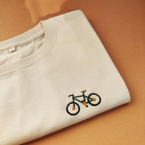 Könnte beinhalten: Ein cremefarbenes T-Shirt mit Rundhalsausschnitt und einer kleinen, gestickten grünen Fahrradgrafik auf der unteren rechten Seite. Das Fahrrad hat schwarze Räder, gelbe Akzente an Pedalen und Rädern sowie einen braunen Sattel. Ein L-Größenetikett ist sichtbar.