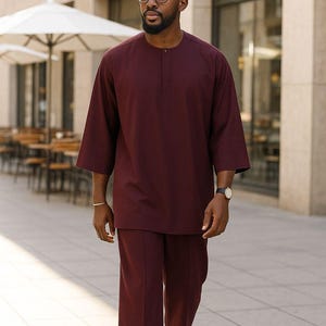 May include: A burgundy two-piece outfit, featuring a long-sleeve tunic and cropped pants. The tunic has a mandarin collar and a buttoned placket. The outfit is paired with black loafers and a silver watch. The ensemble is suitable for formal or semi-formal occasions.